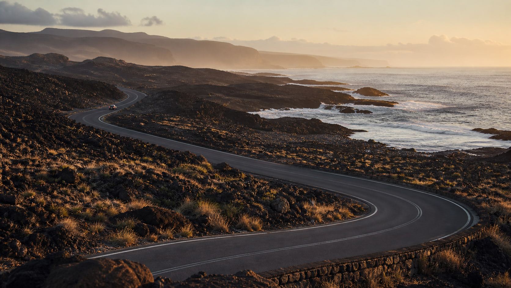 Carretera sinuosa atravesando una costa volcánica al amanecer con un vehículo en la distancia.